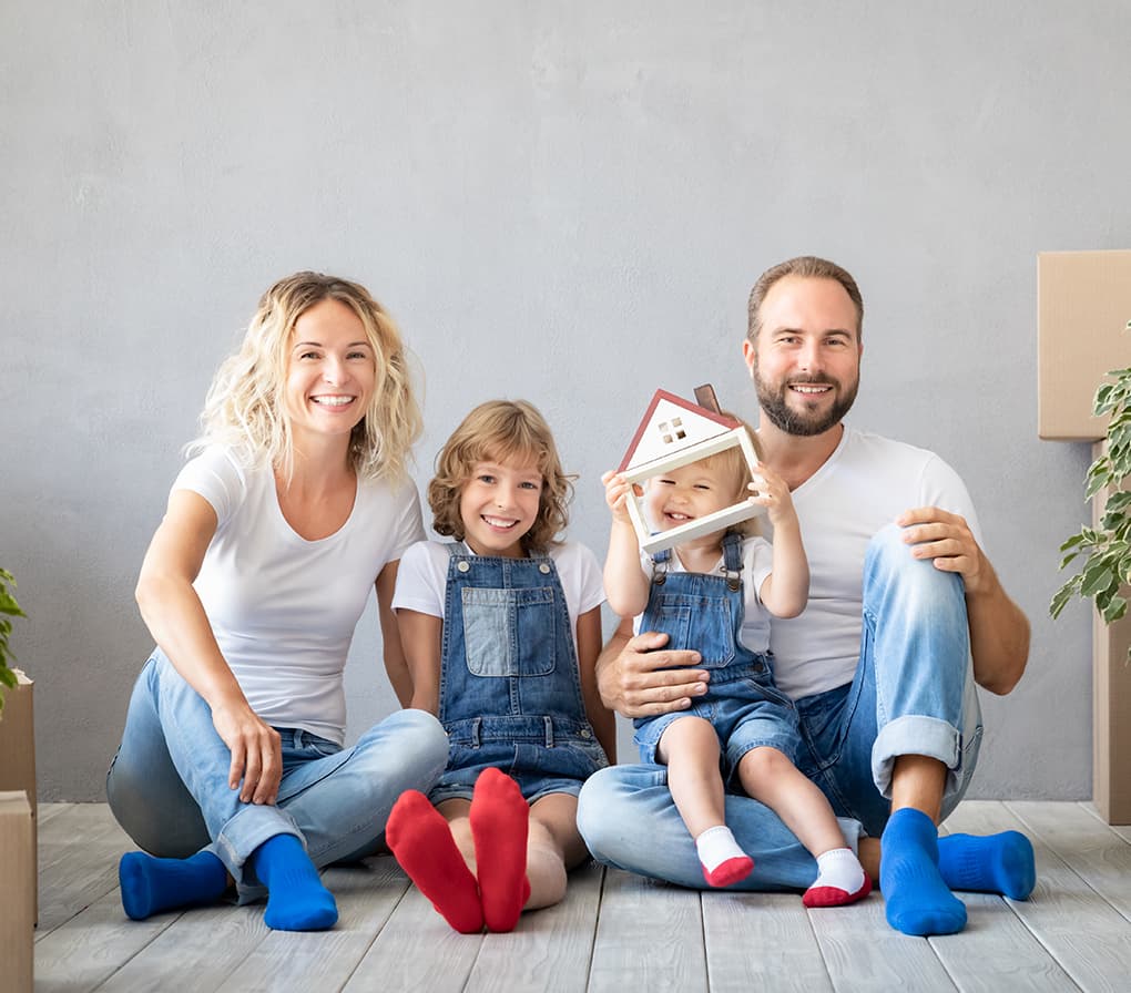 Mosolygó család makettházzal otthonfelújítást ábrázoló jelképes jelenetben, az otthonteremtés, az építkezés, a felújítás és a családiház-építés vizuális megjelenítéseként.