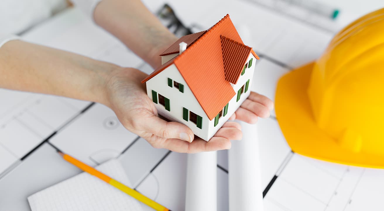 Kézben tartott családi ház makett tervrajzok és védősisak mellett, a kulcsrakész generálkivitelezés jelképeként.