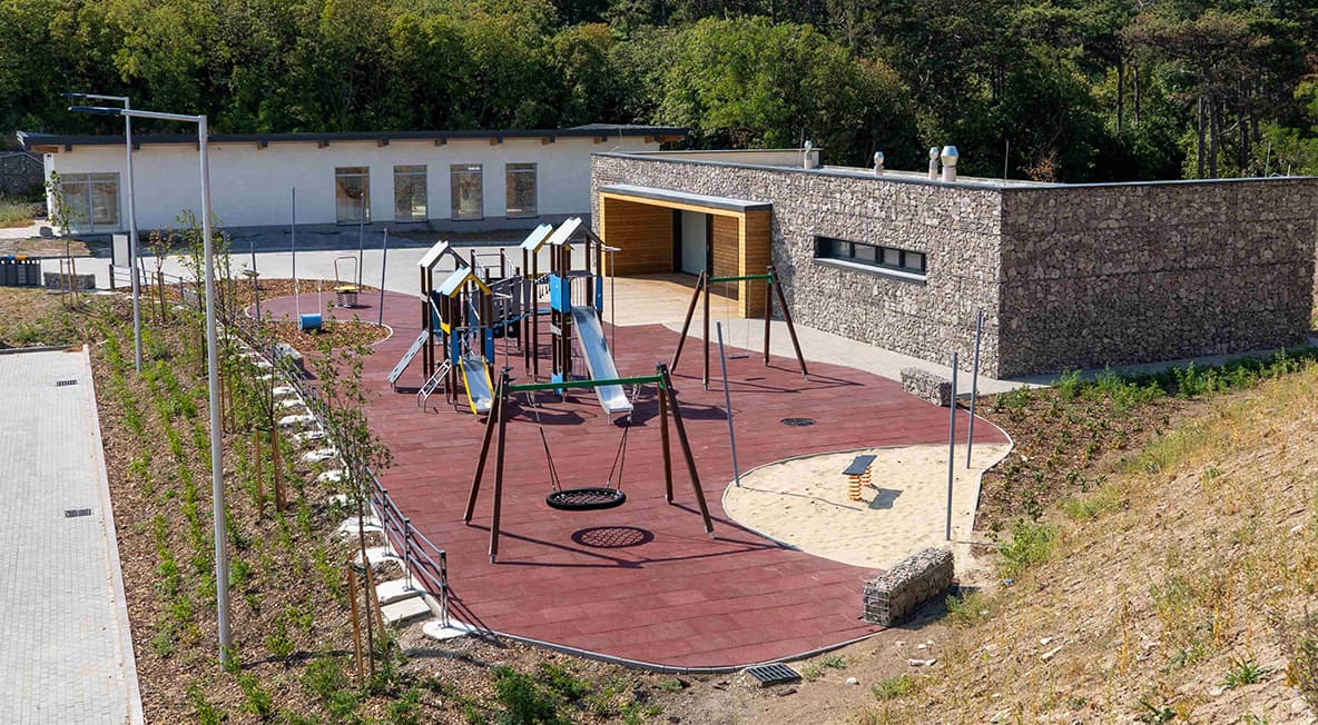 Kiszolgáló épület és játszótér egy rendezett közösségi tér mellett, magasból fotózva.