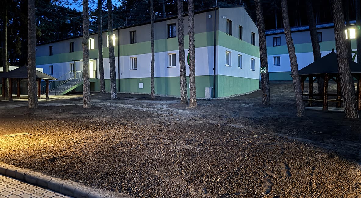 Tábori szállásépület esti megvilágításban, fenyőfák között kialakított környezetben.