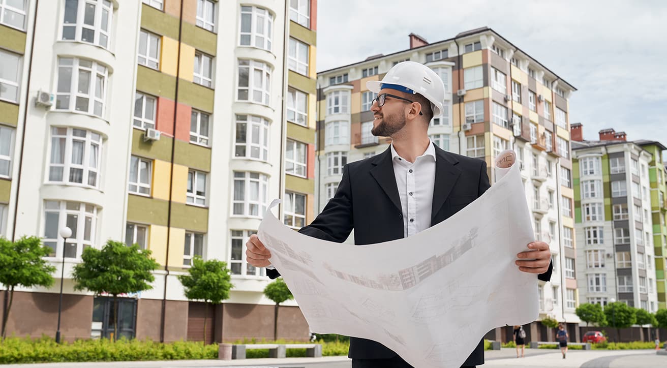 Védősisakos szakember tervrajzot tart társasházak előtt, a társasházkivitelezés tervezési és koordinációs folyamatát szemléltetve.